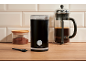 Black coffee grinder, jar of ground coffee, and French press on a wooden countertop.