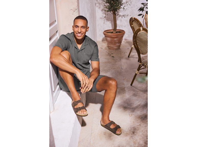 Smiling man in khaki shirt and shorts, brown sandals, sitting on a patio.