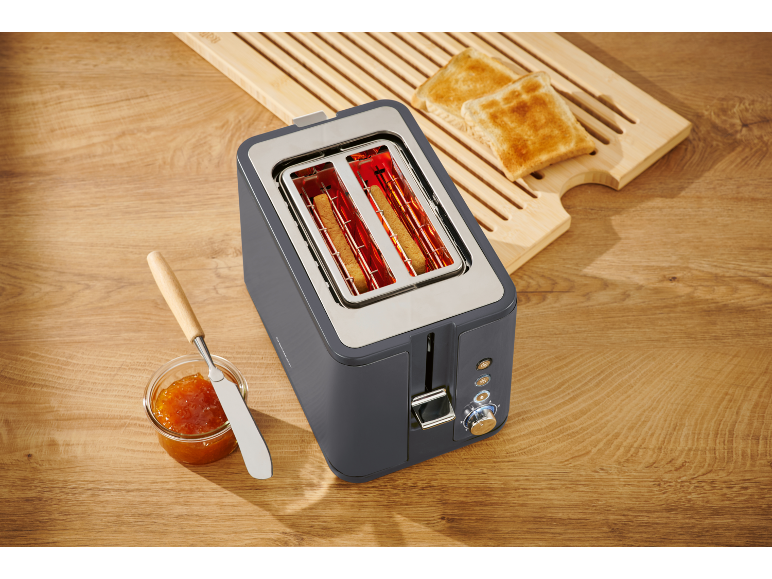 Toaster with bread inside, two slices of toast on a wooden board, and jam with a knife.