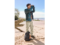 Man in jacket and trousers looking through binoculars on the beach, backpack on the ground.