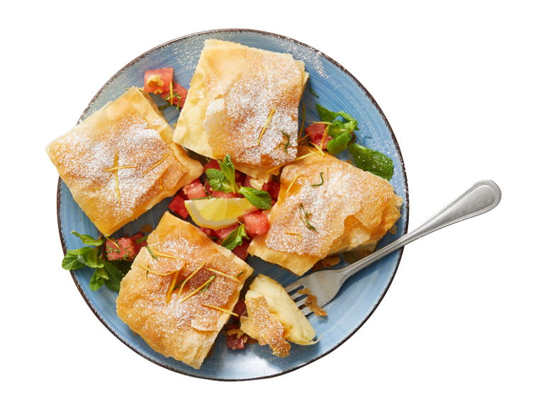 Sweet filo pastry squares with powdered sugar, mint, and watermelon pieces on a blue plate.