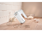 White hand mixer with whisks and dough hooks, baking ingredients on a wooden countertop.