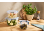 Glass food containers, fresh herbs, and a salt grinder on a wooden countertop.