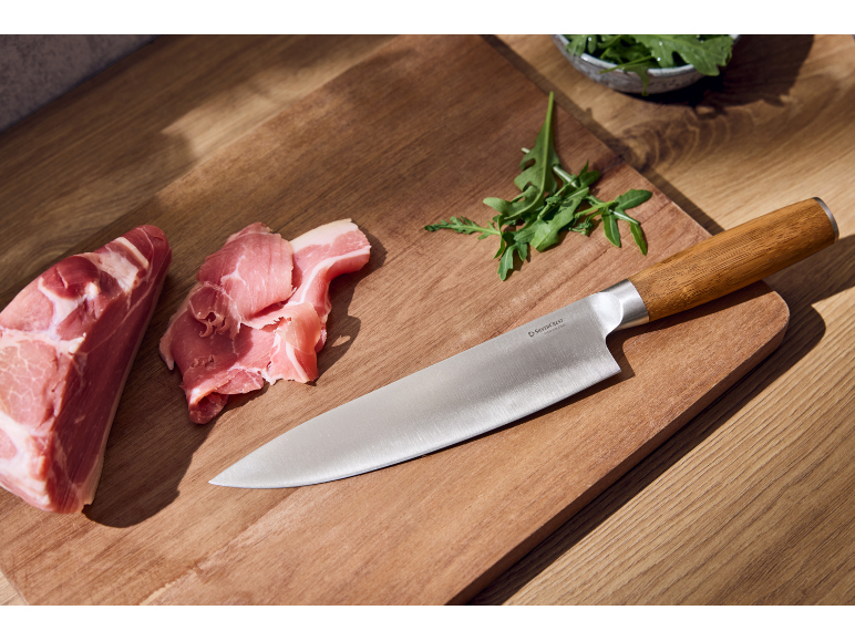 Chef's knife with wooden handle, raw meat, and arugula on a wooden cutting board.