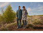 Man and woman in raincoats and boots in nature, with trees and blue sky in the background.