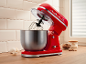 Red kitchen stand mixer with stainless steel bowl, eggs, and baking ingredients.