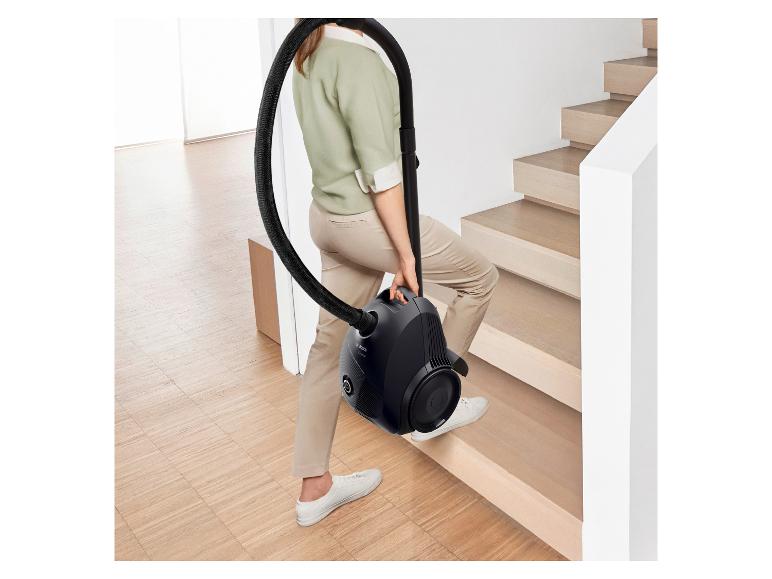 A woman carrying a compact black canister vacuum cleaner up the stairs.