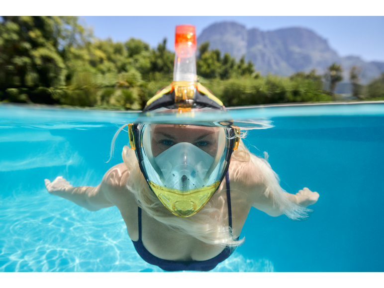 Woman in a full-face snorkel mask swimming in a pool with mountains in the background.