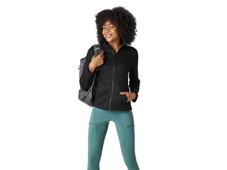 Smiling woman in black jacket and aqua sports leggings, with a gray backpack.