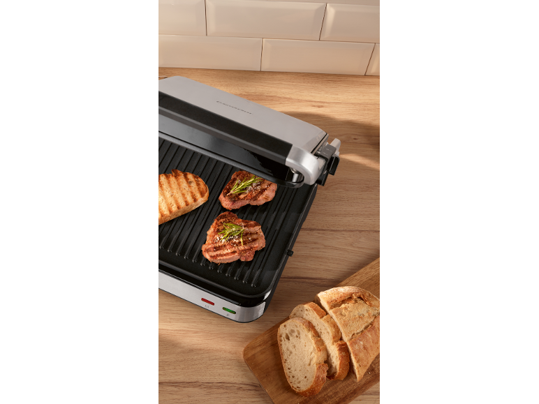 Electric grill with two steaks and a slice of toasted bread, sliced bread on a board.