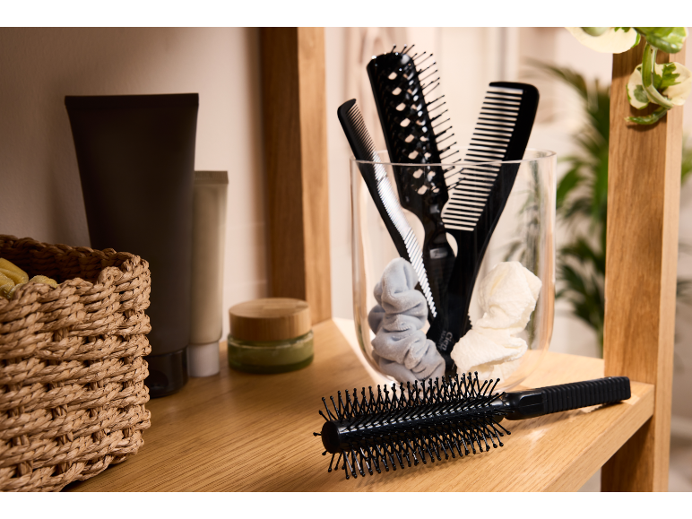 Hairbrushes and combs in a glass, with beauty products and a basket on a wooden shelf.