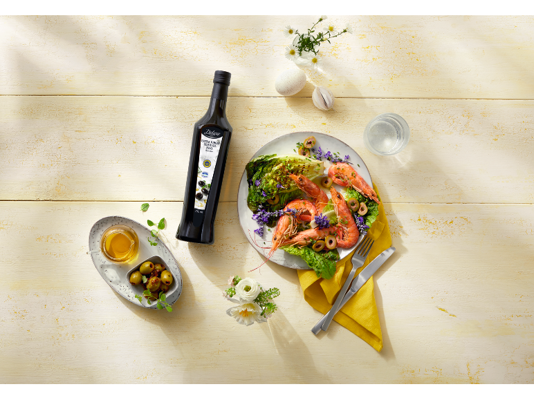 Bottle of olive oil, shrimp salad, and olives on a light wooden table.