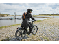 Cyclist in rain jacket and backpack on an e-bike by the water.