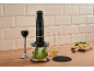 Black hand blender with accessories, herbs, and cheese on a wooden countertop.