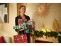 Smiling woman holding a stack of Christmas gifts and a bag with „Merry Christmas” text.