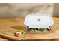 Vacuum food container with salad on a wooden table, next to a cutting board with ingredients.