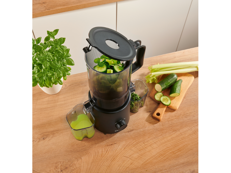 Black juicer on a wooden counter, making green juice from cucumbers and celery.