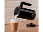 A black milk frother pours foam into a glass latte mug.