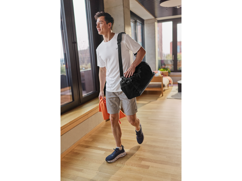 Man in white t-shirt, grey shorts, and sneakers, with a black sports bag and orange towel.