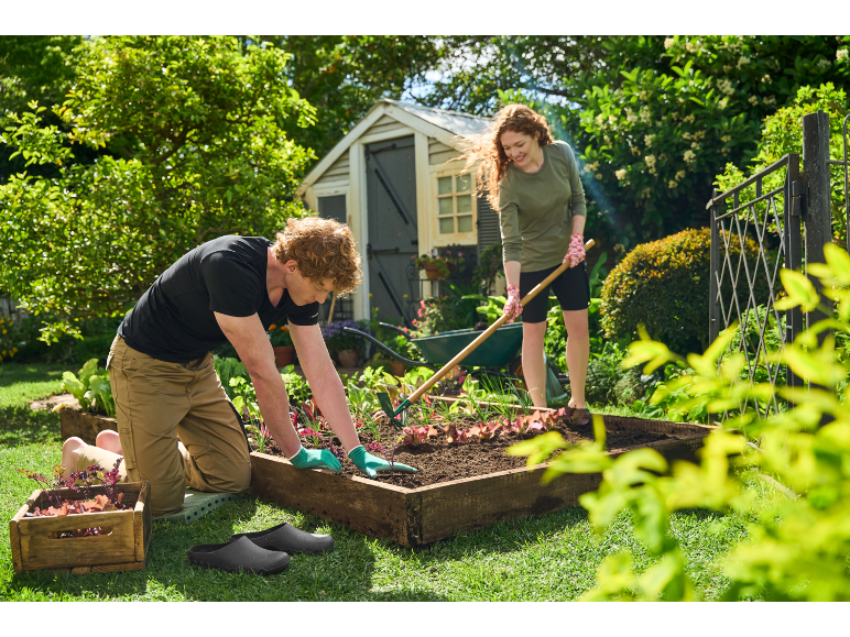 Man and woman planting in a raised garden bed, using gardening tools and gloves.