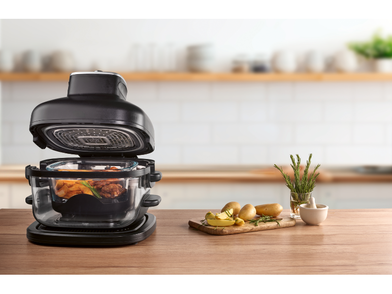 Multicooker with food cooking, potatoes, and rosemary on a wooden countertop.