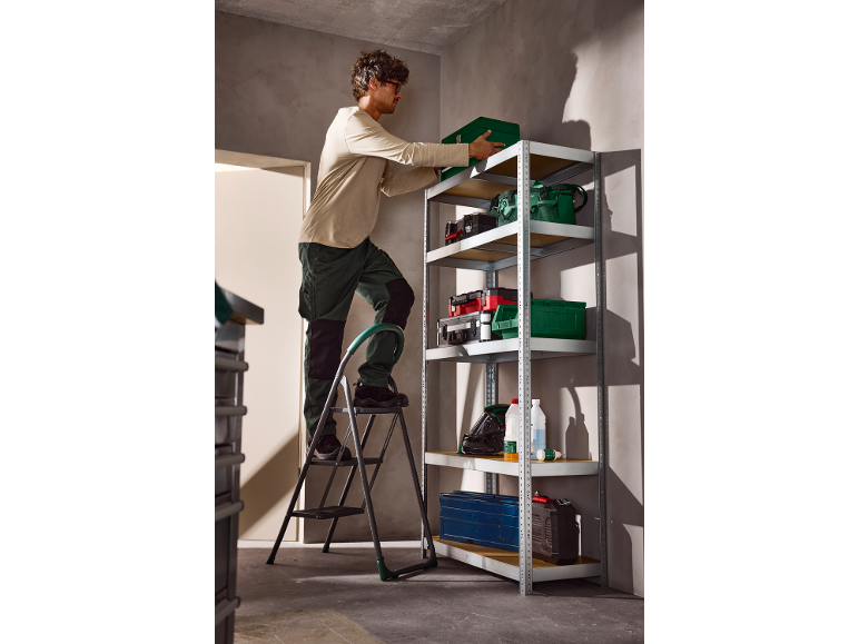 Man on a step ladder placing a green toolbox onto a metal shelving unit.