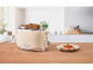 Cream-colored toaster with bread slices and a garnished slice of bread on a wooden table in a kitchen.