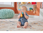 Smiling baby in denim romper, sitting on a rug, playing with a colorful rattle.