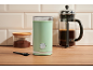 Green coffee grinder, glass jar of ground coffee, and French press on a wooden countertop.