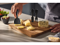 Hand slicing cheese with a cheese slicer on a wooden board with various cheeses and olives.