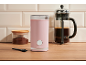 Pink coffee grinder, ground coffee jar, and French press on a wooden table.