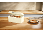 Pretzel and baked goods in a cream-colored bread bin on a wooden kitchen table.