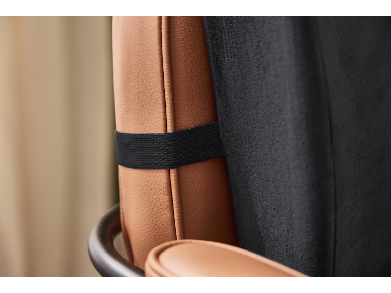 Detail of a brown leather lumbar cushion with a black elastic strap on a chair.