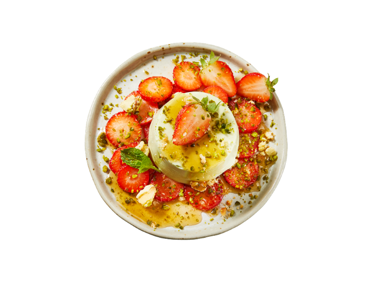 Panna cotta with strawberries, pistachios, and honey on a white plate.