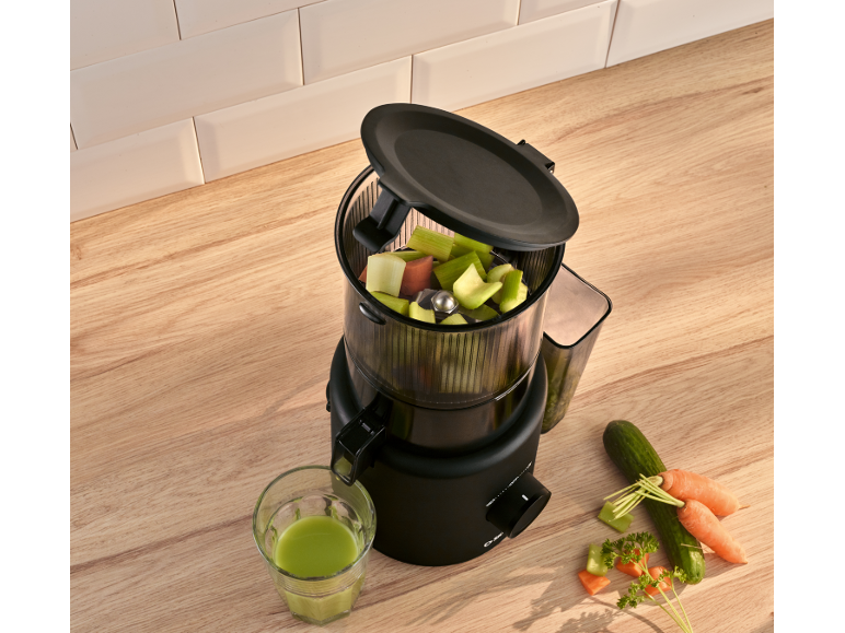 Black juicer with chopped vegetables and a glass of green juice on a wooden countertop.