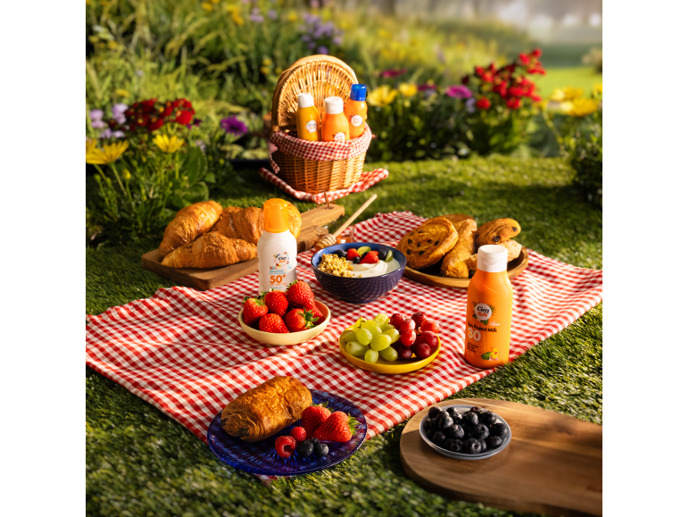 Picnic blanket filled with croissants, fruits, and sunscreens on green grass.