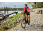 Woman in a red jacket and helmet riding an electric bike on a cobblestone path by a river.