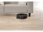 Black robot vacuum cleaner cleaning under a light-colored sofa on a wooden floor.