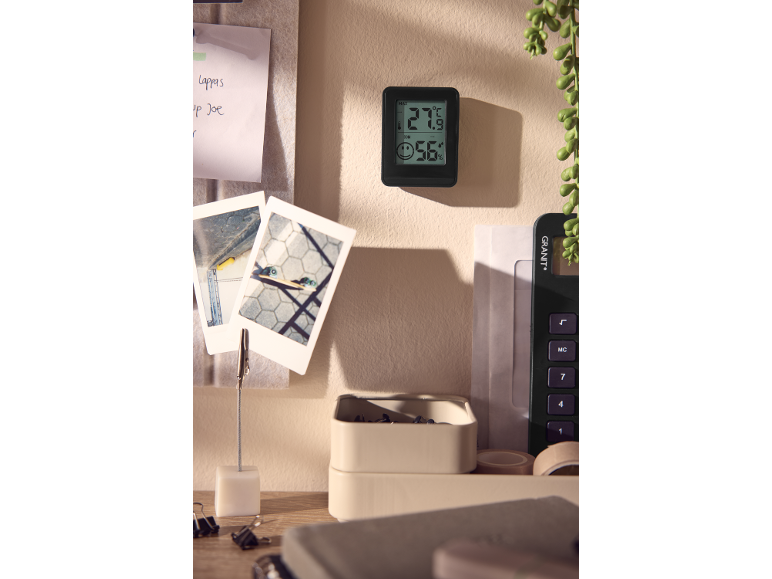 Digital thermometer displaying 27.9°C and 56% humidity on a wall, next to notes and photos.