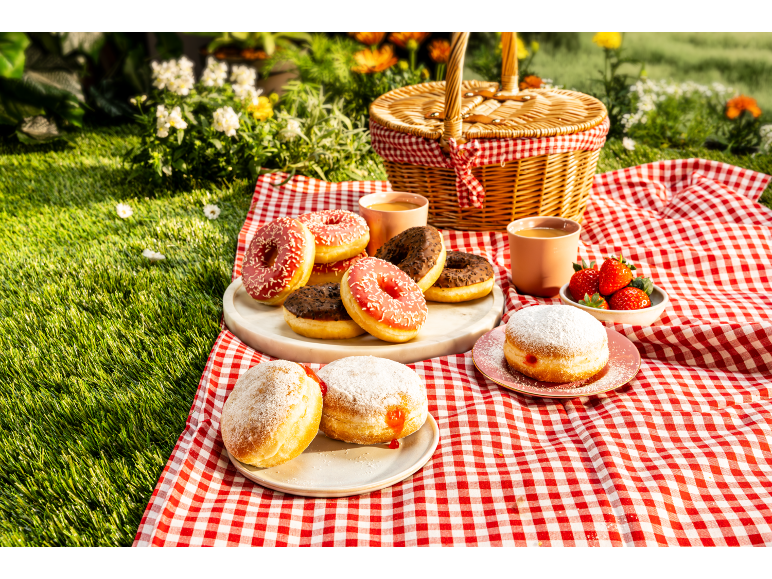 Donuts of various flavors and strawberries at a picnic with a basket and checkered blanket.
