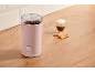 Pink coffee grinder with beans, a glass of coffee, and cookies on a wooden surface.