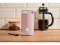 Pink coffee grinder, container with ground coffee, and French press on a wooden surface.