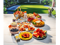 Picnic table with fruits, snacks, juices, and sunscreen in a garden.