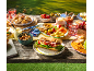 An outdoor picnic with sandwiches, fruit, cheese, crackers, and drinks on a wooden table.