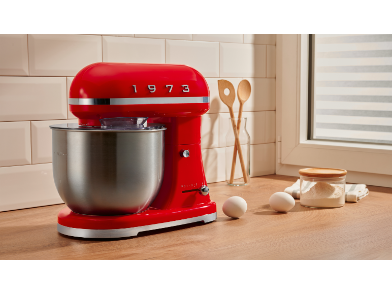 Red stand mixer with stainless steel bowl, eggs, and flour on a wooden countertop.