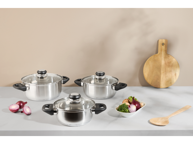 Stainless steel pot set with glass lids, fresh vegetables, and a wooden cutting board.