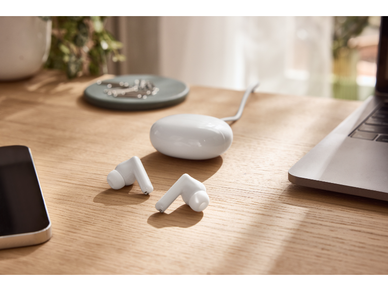 White wireless earbuds and their charging case on a wooden desk with a laptop and phone.