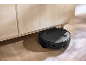Black robot vacuum cleaner cleaning under a light wood cabinet.