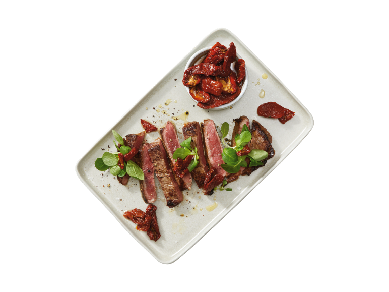 Sliced steak with sun-dried tomatoes and green leaves on a rectangular platter.