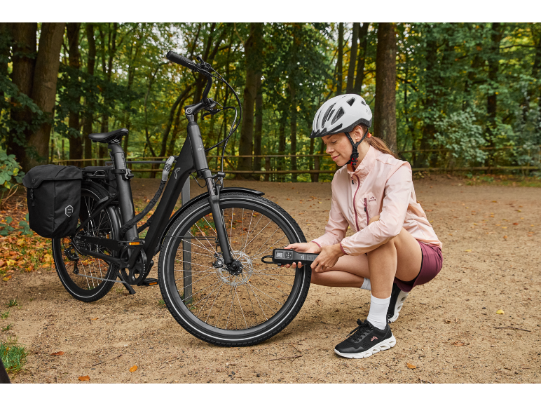 Woman checking tire pressure of an e-bike with a pannier in a forest setting.